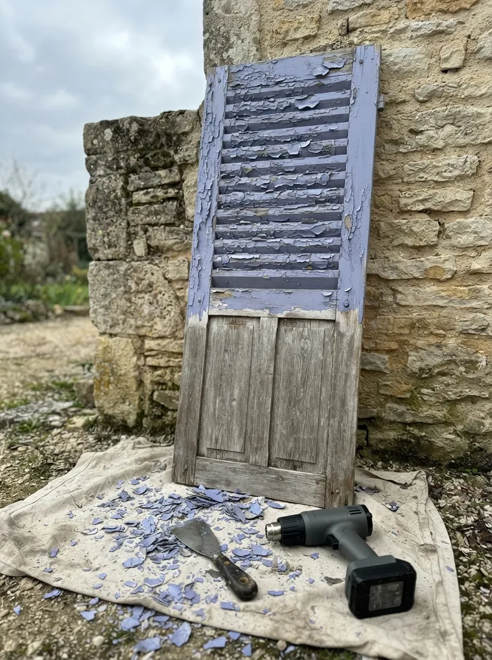 Volet battant en bois appuyé contre un mur en pierre, peinture bleu lavande écaillée en haut, bois nu en bas, décapeur sans fil et grattoir sur une bâche